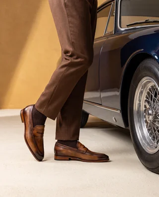 Brown Mastrich penny loafers styled with brown trousers beside a classic car